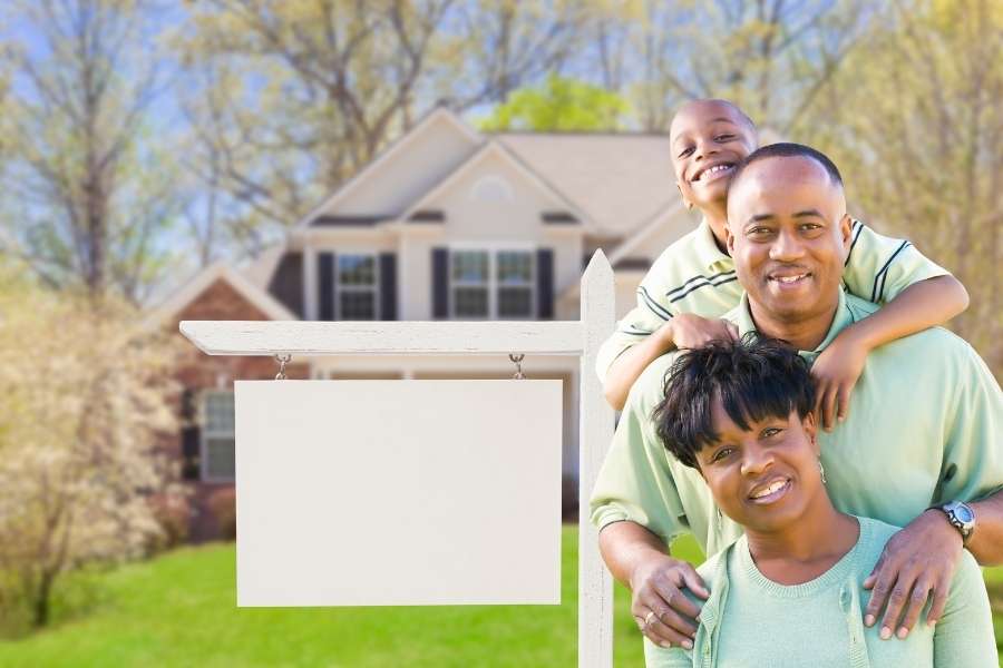 A cheerful family posing in front of their new house with a blank sign representing investment in multi-family properties.