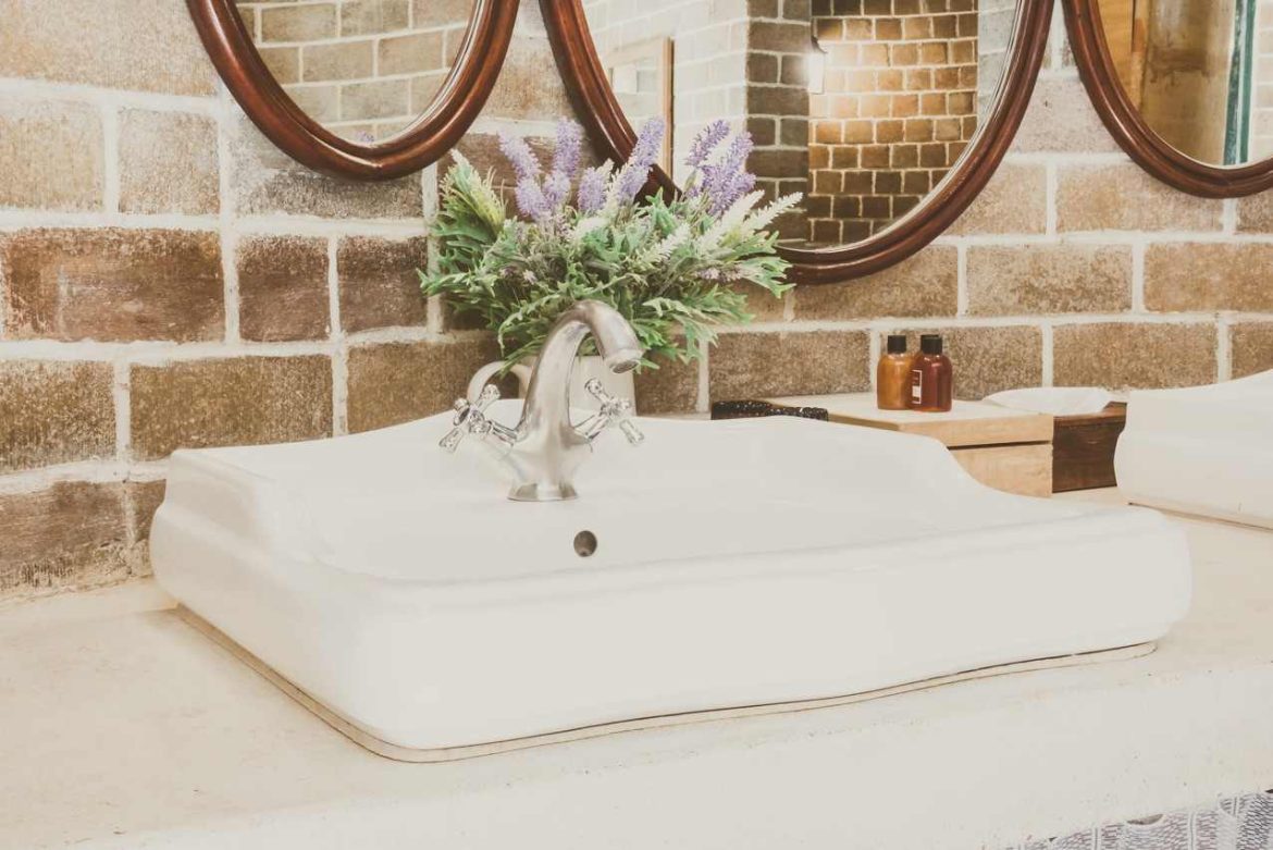 White ceramic basin mounted on a countertop with a classic faucet and brick wall background, illustrating how to choose a bathroom sink.
